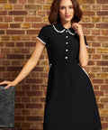 A woman wearing a black short-sleeve A-line ModCloth dress with a white lace trimmed Peter Pan collar and white button details, posing in front of a rustic brick wall.