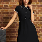 A woman wearing a black short-sleeve A-line ModCloth dress with a white lace trimmed Peter Pan collar and white button details, posing in front of a rustic brick wall.