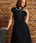Model wearing the Lottie black fit-and-flare dress with white lace-trimmed Peter Pan collar, short sleeves, and flower buttons against a brick wall.