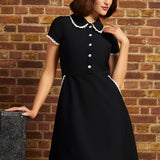 Model wearing the Lottie black fit-and-flare dress with white lace-trimmed Peter Pan collar, short sleeves, and flower buttons against a brick wall.