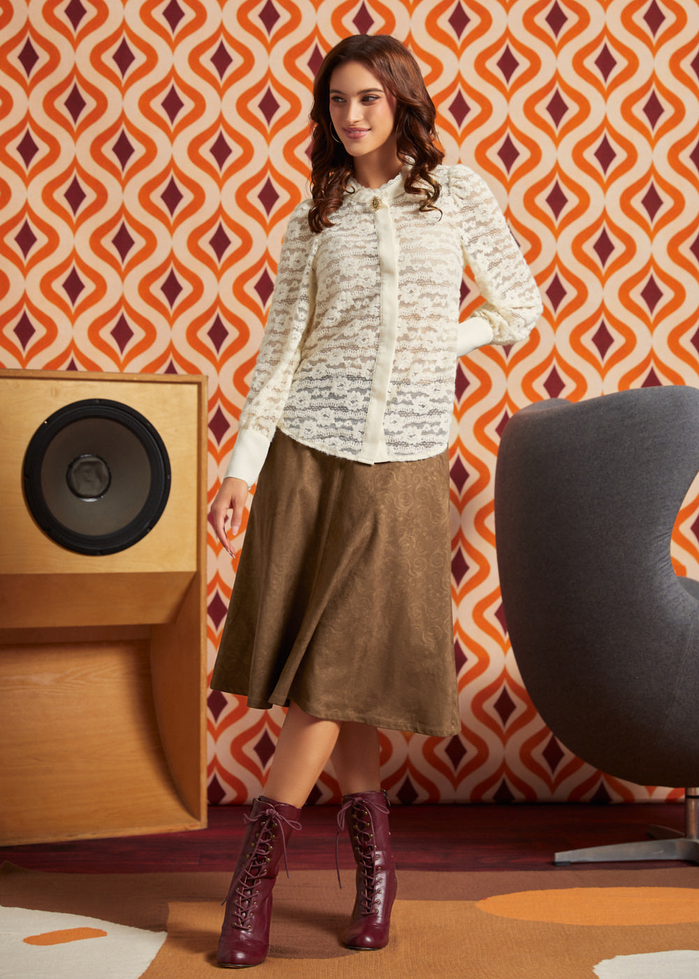 A woman models a long-sleeve white button-up shirt with a sheer floral lace pattern, paired with a brown midi skirt and burgundy lace-up ankle boots, set against a retro orange and white patterned wallpaper background.
