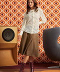 A woman models a long-sleeve white button-up shirt with a sheer floral lace pattern, paired with a brown midi skirt and burgundy lace-up ankle boots, set against a retro orange and white patterned wallpaper background.
