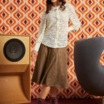 A woman models a long-sleeve white button-up shirt with a sheer floral lace pattern, paired with a brown midi skirt and burgundy lace-up ankle boots, set against a retro orange and white patterned wallpaper background.