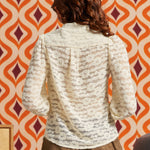 Back view of woman wearing cream floral lace blouse, showing the sheer floral pattern, gathered shoulders, and long puff sleeves.