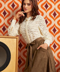 A woman posing in a retro-styled room, wearing a cream-colored textured lace top and a brown skirt, standing in front of a bold orange and burgundy geometric wallpaper and a vintage wooden speaker.