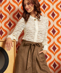 A woman posing in a cream lace long-sleeve button-up blouse and a brown high-waisted skirt with a tie waist, set against a retro orange and white patterned wallpaper background.