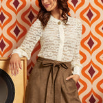 A woman posing in a cream lace long-sleeve button-up blouse and a brown high-waisted skirt with a tie waist, set against a retro orange and white patterned wallpaper background.