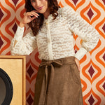 A woman models a white lace long-sleeve blouse and a brown patterned skirt against a retro orange and burgundy patterned wallpaper background.