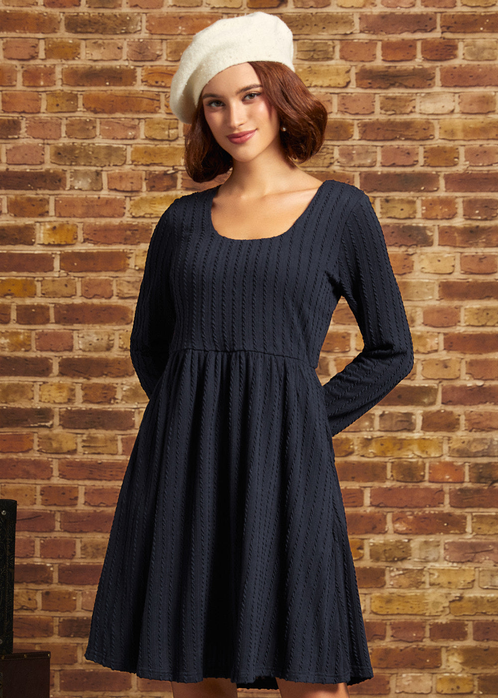 A woman wearing a dark navy blue textured long-sleeve dress and a cream-colored beret, standing against a rustic brick wall background.