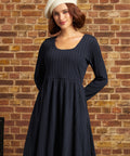 A woman wearing a dark navy blue textured long-sleeve dress and a cream-colored beret, standing against a rustic brick wall background.