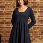 A woman wearing a dark navy blue textured long-sleeve dress and a cream-colored beret, standing against a rustic brick wall background.