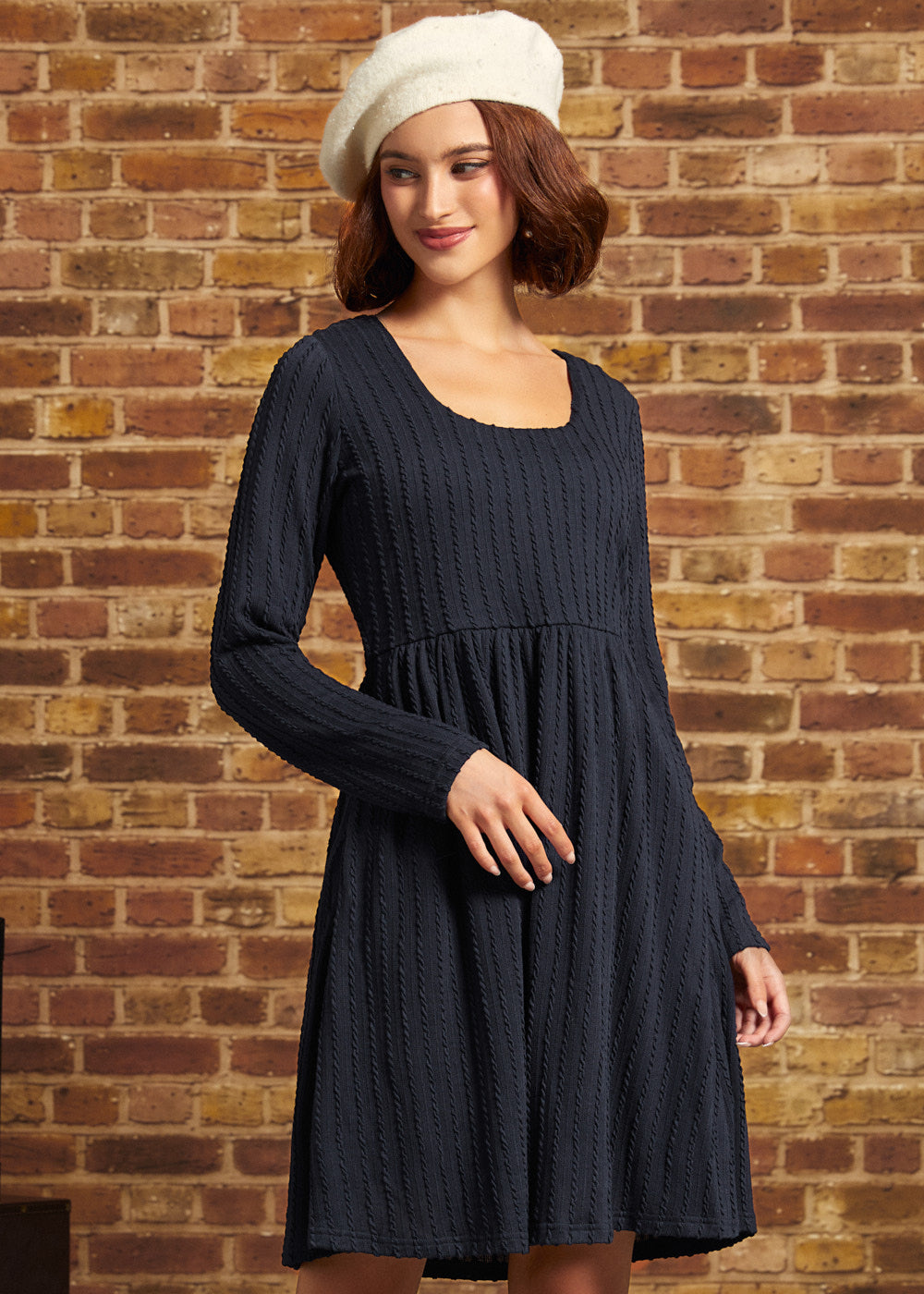 A woman wearing a dark navy blue ribbed knit long-sleeve dress and a cream-colored beret, standing against a rustic brick wall background.