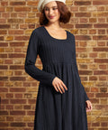 A woman wearing a dark navy blue ribbed knit long-sleeve dress and a cream-colored beret, standing against a rustic brick wall background.