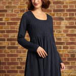 A woman wearing a dark navy blue ribbed knit long-sleeve dress and a cream-colored beret, standing against a rustic brick wall background.