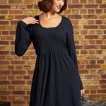 A woman wearing a dark navy blue cable-knit dress and a cream-colored beret, posing in front of a rustic brick wall.