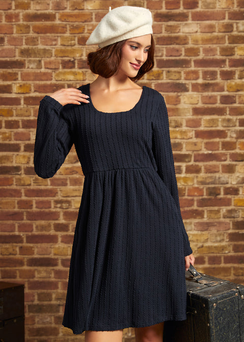 A woman wearing a dark navy blue cable-knit dress and a cream-colored beret, posing in front of a rustic brick wall.