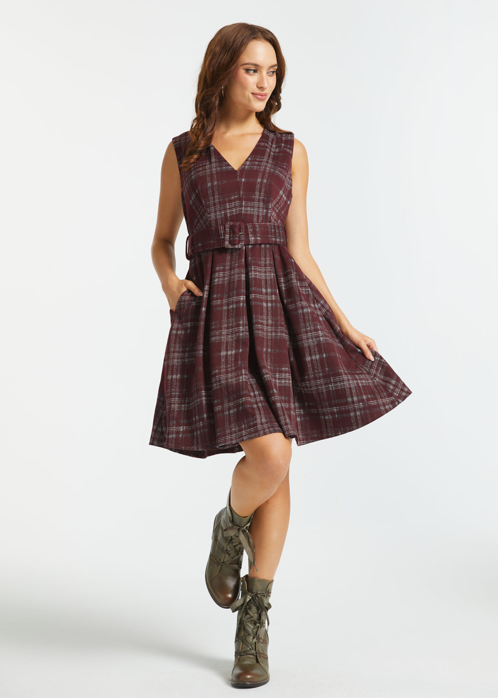A woman modeling a sleeveless plaid dress with a V-neck, waist tie, and flared skirt, paired with brown lace-up ankle boots, against a light gray studio background.