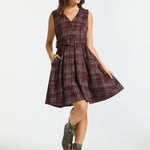 A woman modeling a sleeveless plaid dress with a V-neck, waist tie, and flared skirt, paired with brown lace-up ankle boots, against a light gray studio background.