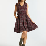 A woman modeling a sleeveless plaid dress with a V-neck, waist tie, and flared skirt, paired with brown lace-up ankle boots, against a light gray studio background.