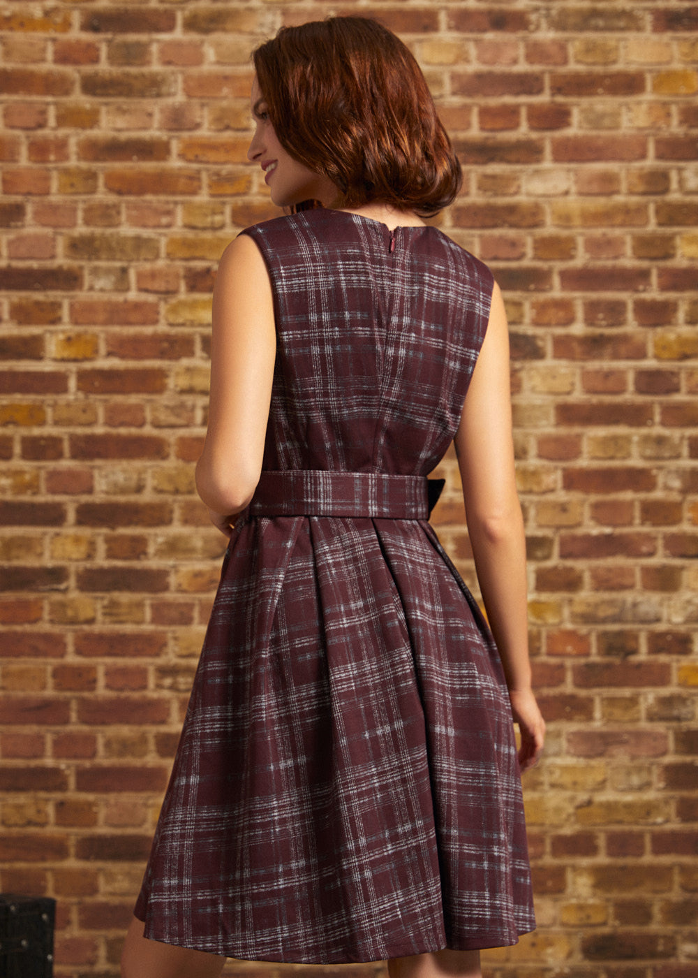 Woman wearing a burgundy plaid sleeveless fit and flare dress, showing the back zipper, matching waist belt, and pleated skirt against a brick wall.