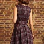 Woman wearing a burgundy plaid sleeveless fit and flare dress, showing the back zipper, matching waist belt, and pleated skirt against a brick wall.