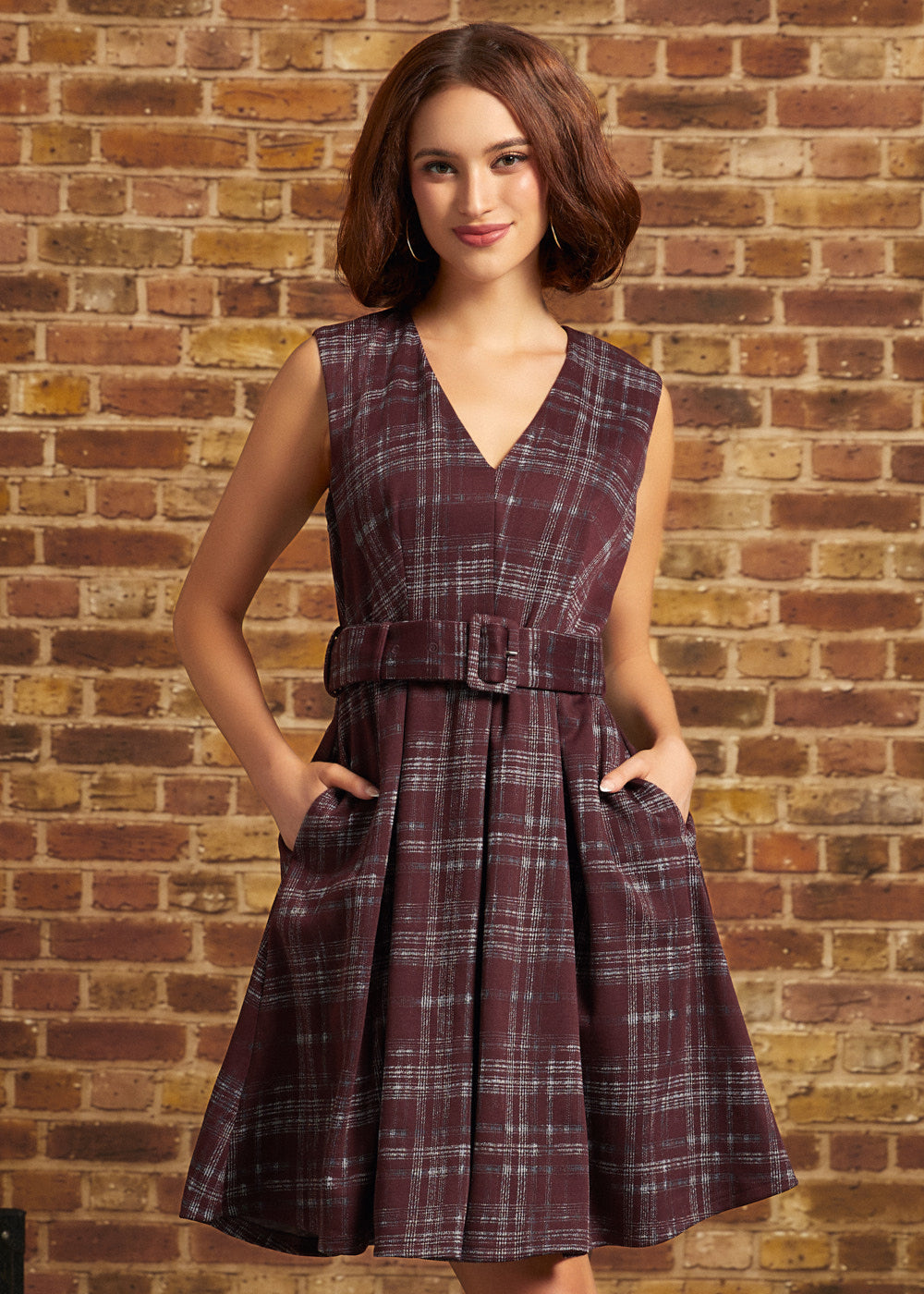 A woman wearing a sleeveless V-neck plaid dress with a matching waist belt, standing against a brick wall.