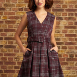 A woman wearing a sleeveless V-neck plaid dress with a matching waist belt, standing against a brick wall.