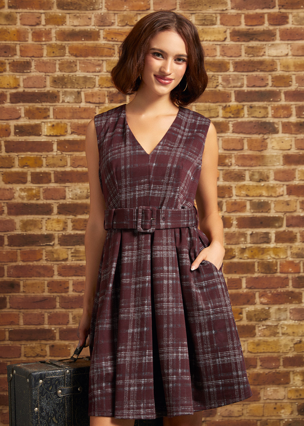 Woman wearing a burgundy plaid sleeveless fit and flare dress with a V-neckline, removable buckle belt, and pleated skirt, standing beside a vintage suitcase against a brick wall.
