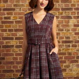 Woman wearing a burgundy plaid sleeveless fit and flare dress with a V-neckline, removable buckle belt, and pleated skirt, standing beside a vintage suitcase against a brick wall.