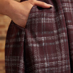 Close-up of a woman's hand tucked into the side pocket of a burgundy plaid pleated dress, showing the fabric texture and gathered skirt folds.