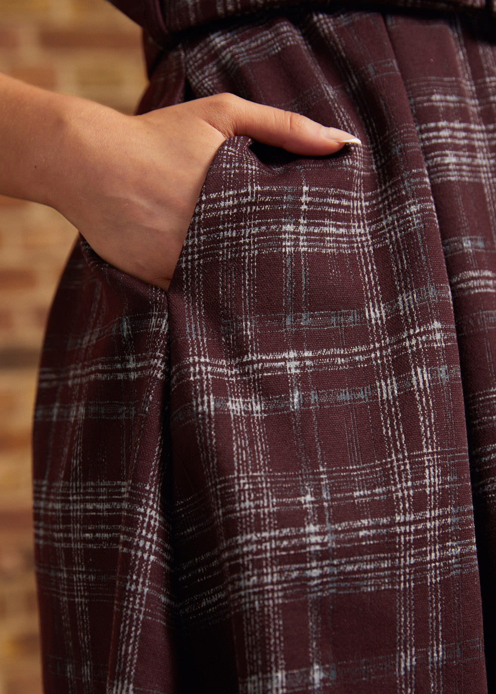 Close-up of a woman's hand tucked into the side pocket of a burgundy plaid pleated dress, showing the fabric texture and gathered skirt folds.