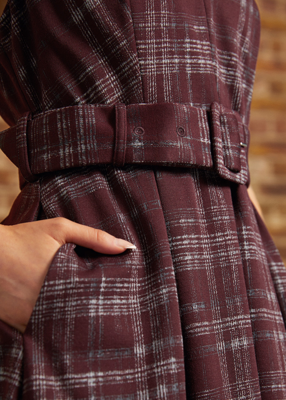 Close-up of a plaid belted dress in deep burgundy with white and gray windowpane lines, showing a hand tucked into a side pocket.
