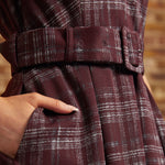 Close-up of a plaid belted dress in deep burgundy with white and gray windowpane lines, showing a hand tucked into a side pocket.