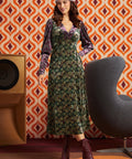 A woman models a 1970s-style midi dress with a green and gold floral pattern, purple velvet long sleeves, and a matching purple headband, standing in front of a retro orange and white patterned wallpaper background.