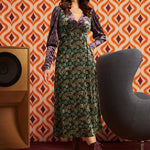 A woman models a 1970s-style midi dress with a green and gold floral pattern, purple velvet long sleeves, and a matching purple headband, standing in front of a retro orange and white patterned wallpaper background.