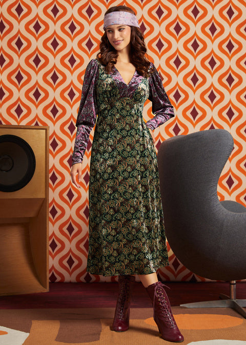 A woman models a 1970s-style midi dress with a green and gold floral pattern, purple velvet long sleeves, and a matching purple headband, standing in front of a retro orange and white patterned wallpaper background.