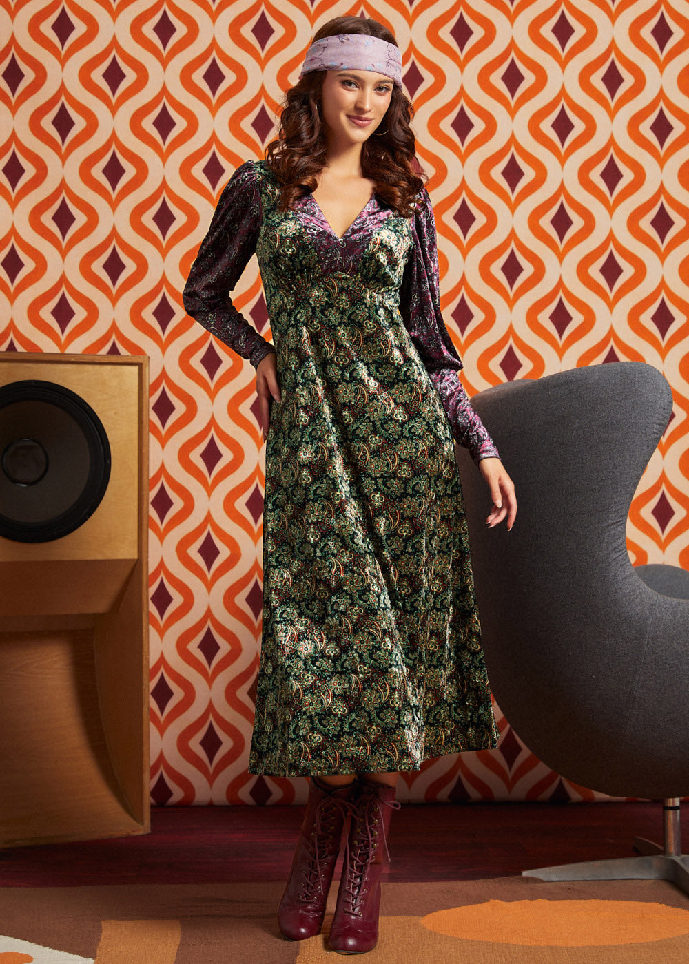 A woman models a long-sleeved midi dress with a green and gold floral pattern and purple sleeves, styled with a purple headband and burgundy lace-up boots, set against a retro orange and brown patterned wallpaper background.