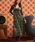 A woman models a long-sleeved midi dress with a green and gold floral pattern and purple sleeves, styled with a purple headband and burgundy lace-up boots, set against a retro orange and brown patterned wallpaper background.