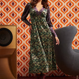 A woman models a long-sleeved midi dress with a green and gold floral pattern and purple sleeves, styled with a purple headband and burgundy lace-up boots, set against a retro orange and brown patterned wallpaper background.