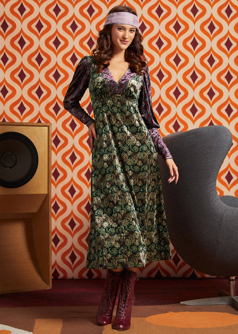 A woman models a long-sleeved midi dress with a green and gold floral pattern and purple sleeves, styled with a purple headband and burgundy lace-up boots, set against a retro orange and brown patterned wallpaper background.