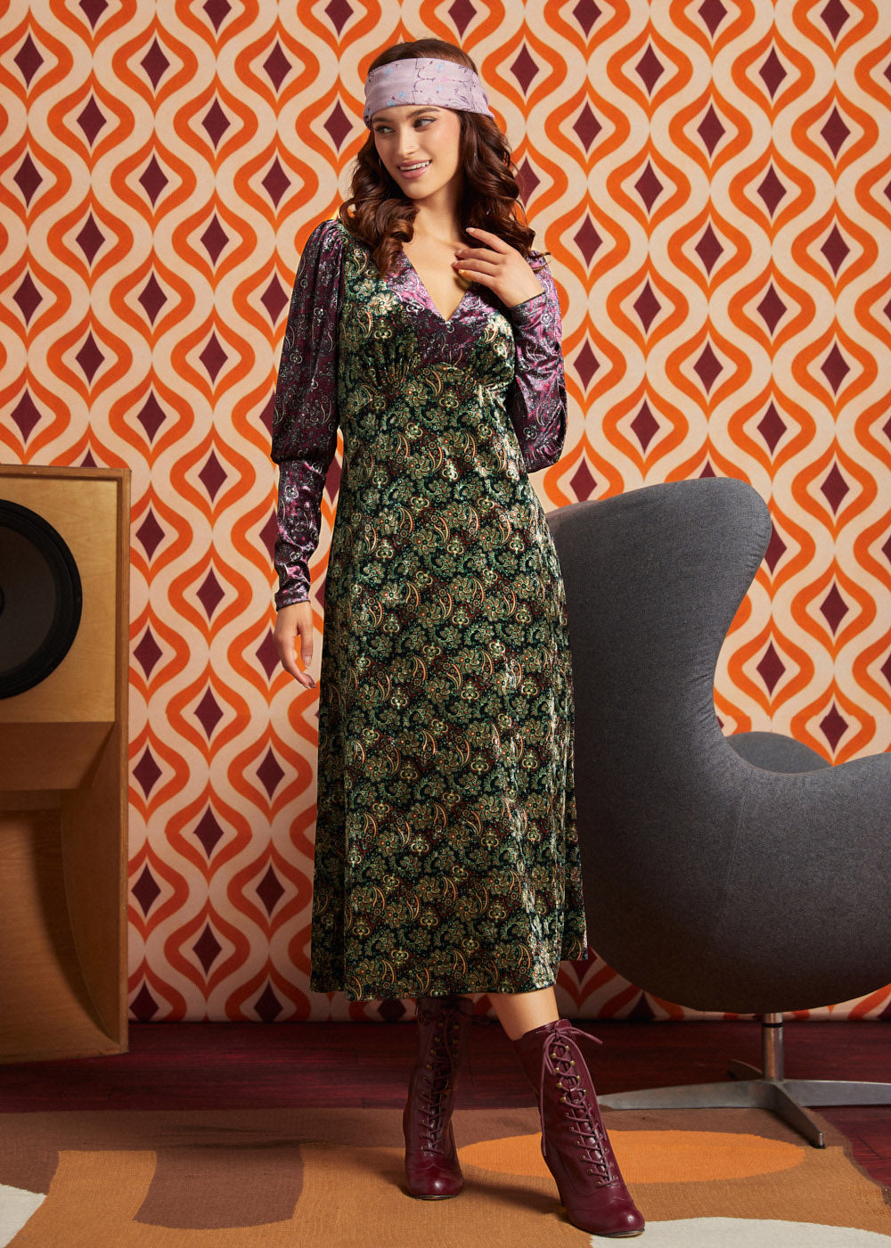 A woman models a 1970s-style ModCloth green floral midi dress with a purple velvet long-sleeve layer and matching headband, standing in front of retro orange and white patterned wallpaper.