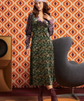 A woman models a 1970s-style ModCloth green floral midi dress with a purple velvet long-sleeve layer and matching headband, standing in front of retro orange and white patterned wallpaper.