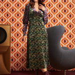 A woman models a 1970s-style ModCloth green floral midi dress with a purple velvet long-sleeve layer and matching headband, standing in front of retro orange and white patterned wallpaper.