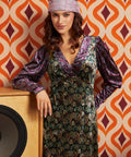 A woman wearing a long-sleeved, V-neck dress with a purple and green paisley pattern and a matching headband, posing in front of a retro orange and brown patterned wallpaper background.