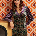 A woman wearing a long-sleeved, V-neck dress with a purple and green paisley pattern and a matching headband, posing in front of a retro orange and brown patterned wallpaper background.