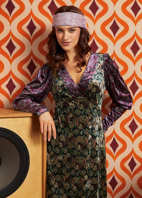 A woman wearing a long-sleeved, V-neck dress with a purple and green paisley pattern and a matching headband, posing in front of a retro orange and brown patterned wallpaper background.