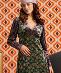 A woman wearing a long-sleeved velvet dress with a green and purple paisley pattern, a matching purple patterned headband, and posing in front of a retro orange and white patterned wallpaper background.