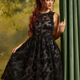 A woman modeling a sleeveless black lace and sequin A-line dress with a high neckline, standing in front of a green curtain and a forest backdrop.