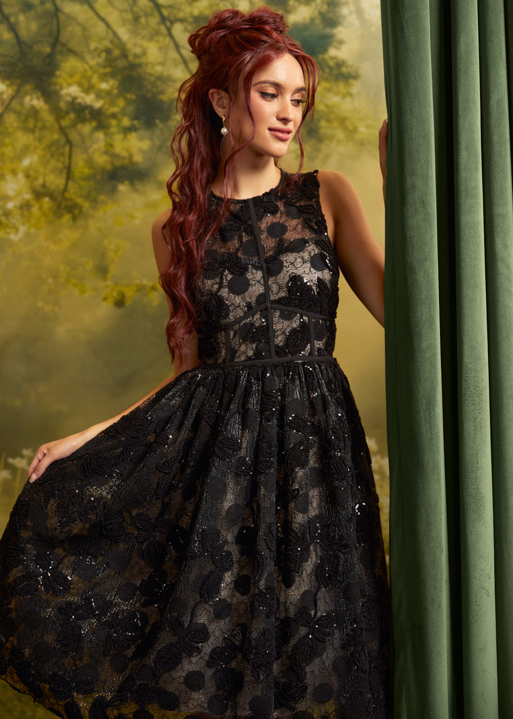 A woman with long red hair wearing a sleeveless black lace dress with a sheer floral pattern and a flared skirt, posed beside a green curtain against a soft yellow-green forest backdrop.