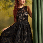 A woman with long red hair wearing a sleeveless black lace dress with a sheer floral pattern and a flared skirt, posed beside a green curtain against a soft yellow-green forest backdrop.
